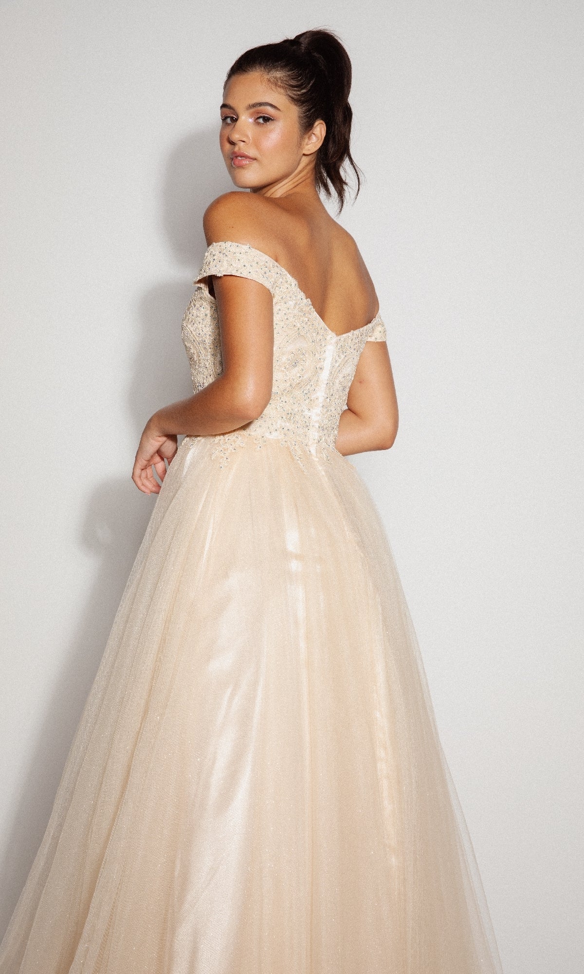 A woman poses against a plain light background in the Off-Shoulder Glitter Prom Ball Gown: Eureka 9899, featuring a fitted, embellished bodice and a flowing tulle skirt.